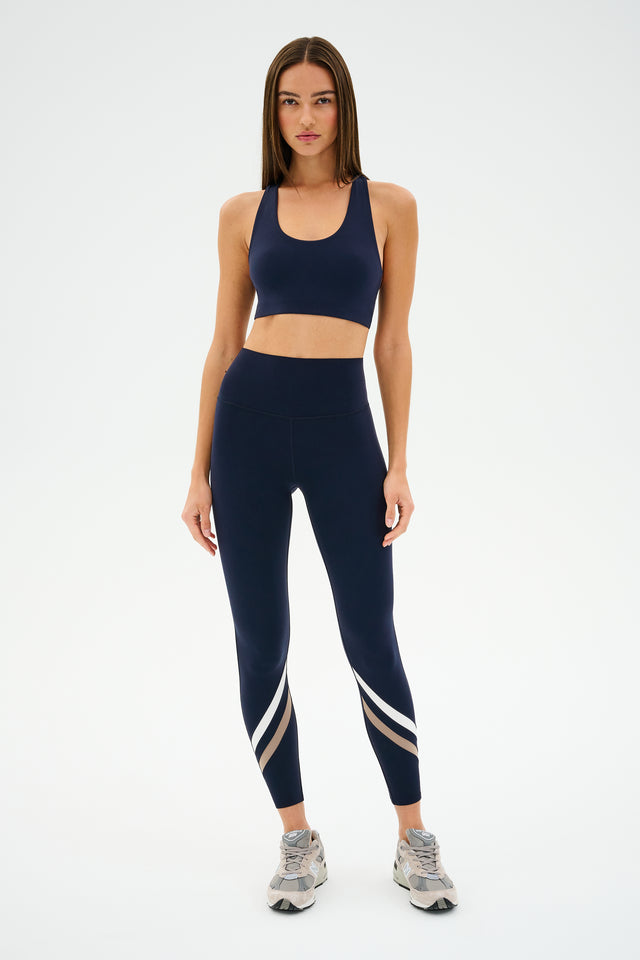 A person in navy athletic wear stands against a plain background, donning SPLITS59's Chevron Airweight High Waist 7/8 leggings in Indigo/Latte, paired with a sports bra and sleek sneakers.