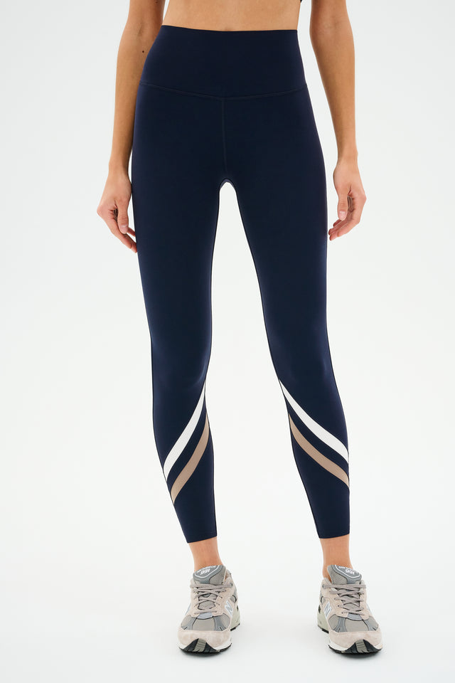 A person in SPLITS59's Chevron Airweight High Waist 7/8 Indigo/Latte leggings and gray sneakers stands against a plain background, highlighting the luxe fabric that effortlessly blends style and comfort.