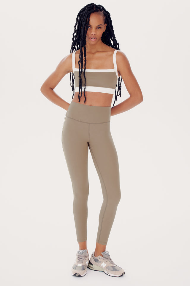 Woman in SPLITS59 Monah Rigor Bra - Latte/White and olive green activewear, standing in a studio with a retro sport vibe, and long braided hair.