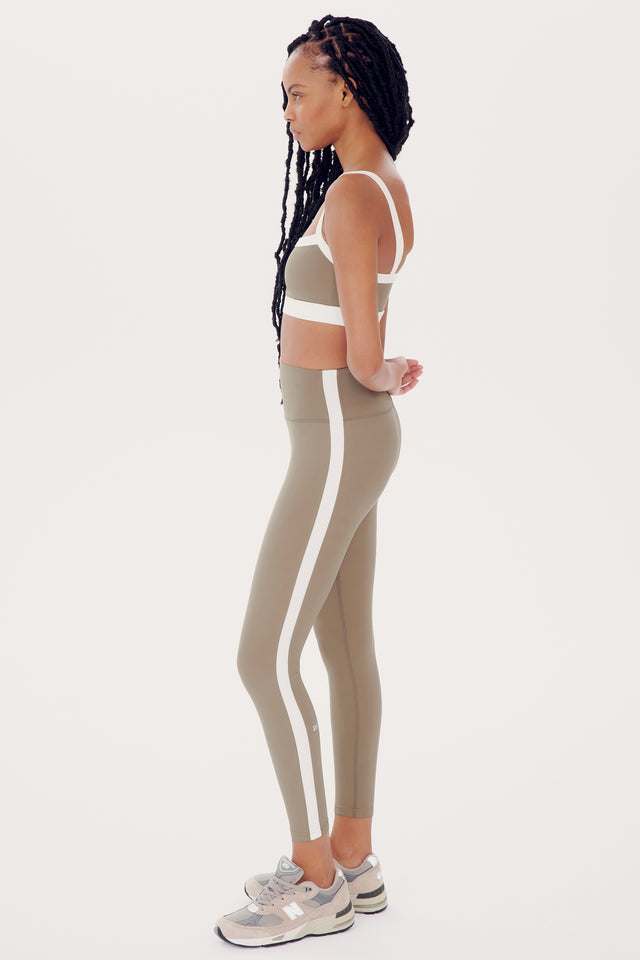 A woman in profile wearing a white sports bra and olive Clare High Waist Rigor 7/8 leggings with a white stripe, standing against a white background by SPLITS59.