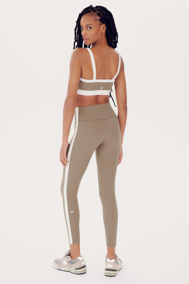 A woman in athletic wear, including a SPLITS59 Monah Rigor Bra in Latte/White and olive leggings with white stripes, stands in a studio and looks over her shoulder, channeling a retro sport vibe.