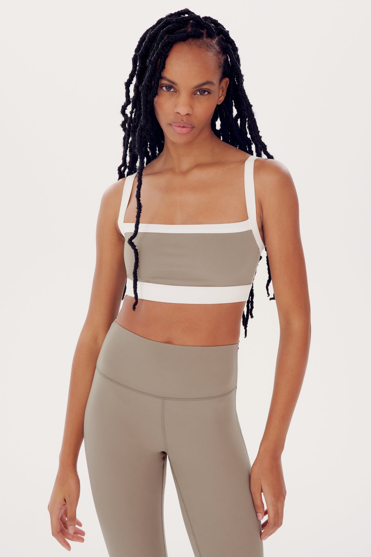 A woman with long braided hair wearing a SPLITS59 Monah Rigor Bra in Latte/White and beige leggings, standing against a white background.