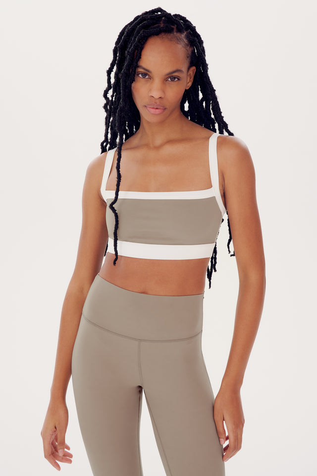 A woman with long braided hair wearing a SPLITS59 Monah Rigor Bra in Latte/White and beige leggings, standing against a white background.
