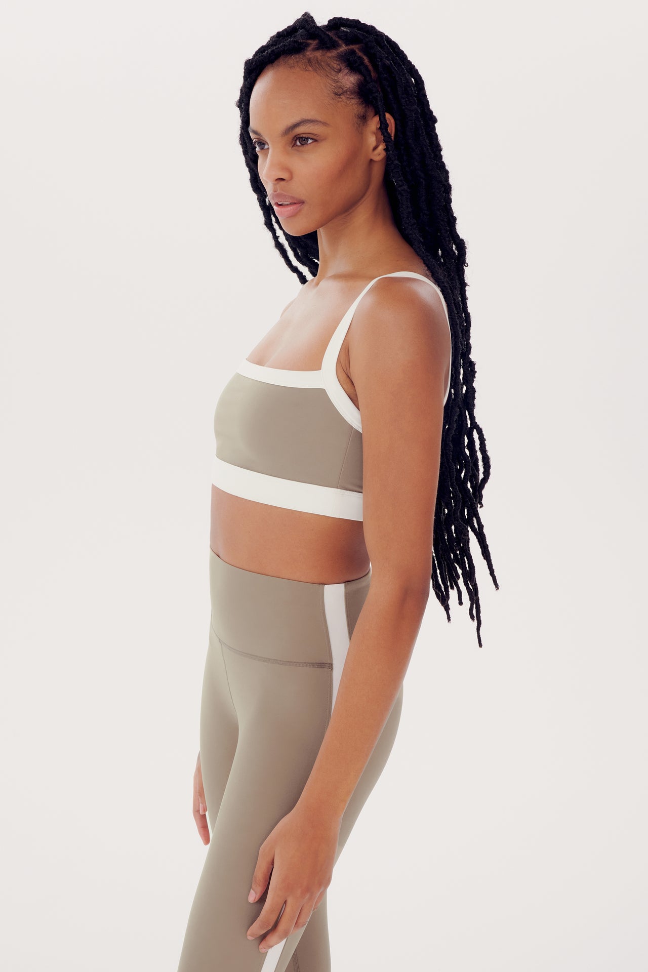 A woman with long braided hair wearing a SPLITS59 Monah Rigor Bra in Latte/White and leggings stands in a profile pose against a white background.