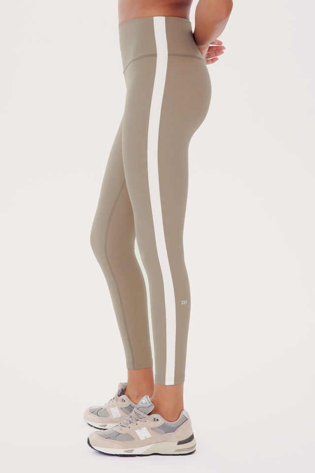 Side view of a woman in SPLITS59 Clare High Waist Rigor 7/8 - Latte/White high waist leggings with white side stripes, standing and showcasing the fit, paired with light grey sneakers.
