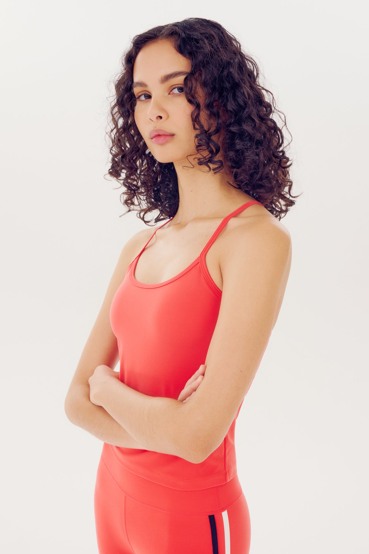 A woman with curly hair wearing a red athletic Airweight Tank by SPLITS59, made of nylon spandex fabric, standing with crossed arms against a white background.