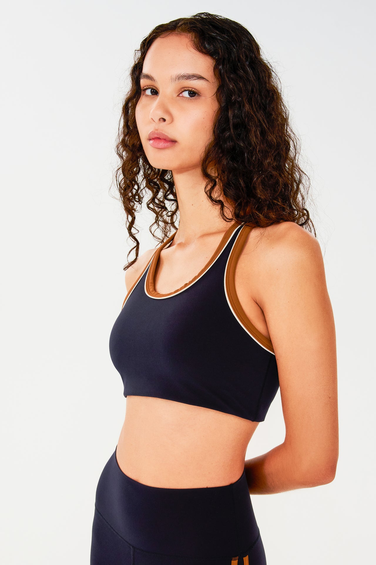 Front side view of woman with dark wavy hair wearing dark blue bra with white and dark orange border arm and neckline and racerback and dark blue leggings with dark orange double side stripes