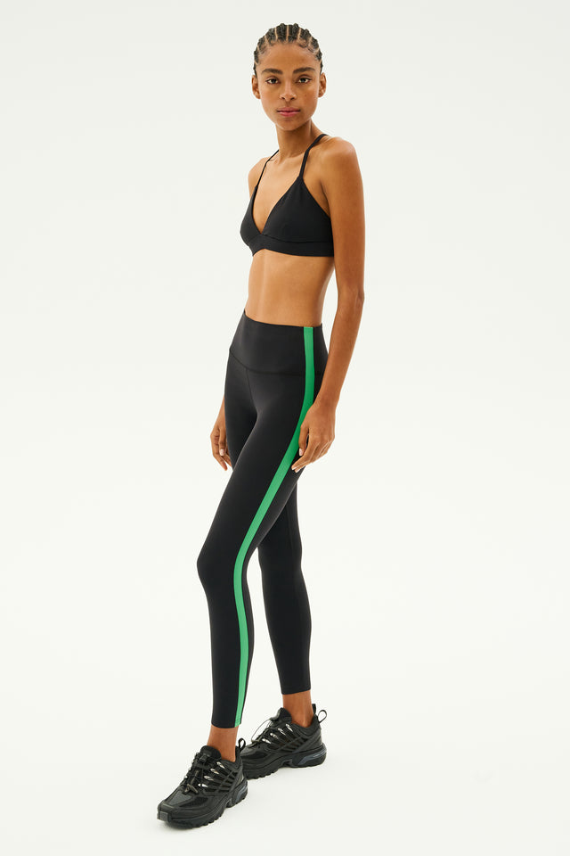 Dressed in a black sports bra and SPLITS59's Clare High Waist Rigor 7/8 leggings in Black/Grass, expertly designed with high-performance fabric, and paired with black sneakers, a person stands poised for gym workouts against a plain white background.