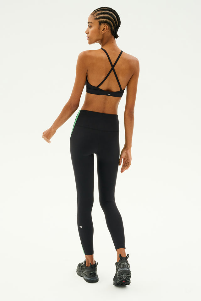 Against a light background, the individual is wearing a black sports bra along with SPLITS59's Clare High Waist Rigor 7/8 leggings in Black/Grass, featuring a sleek green stripe. Ideal for gym workouts, these leggings are crafted from high-performance fabric and are paired with stylish black sneakers.