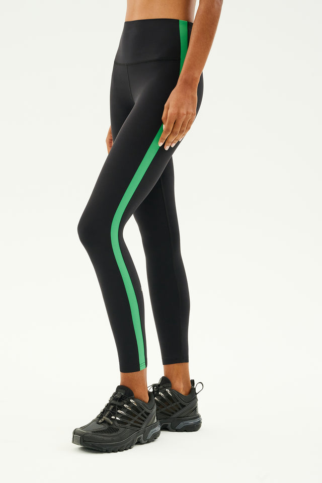 A person wearing the SPLITS59 Clare High Waist Rigor 7/8 in Black/Grass and black sneakers stands against a plain background, highlighting the style and comfort of the high-performance Rigor fabric.