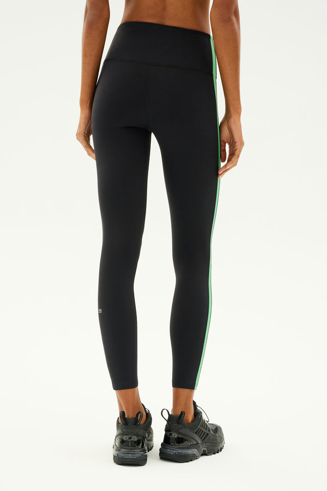 A person wearing SPLITS59's Clare High Waist Rigor 7/8 leggings in Black/Grass and black sneakers stands with their back to the camera.