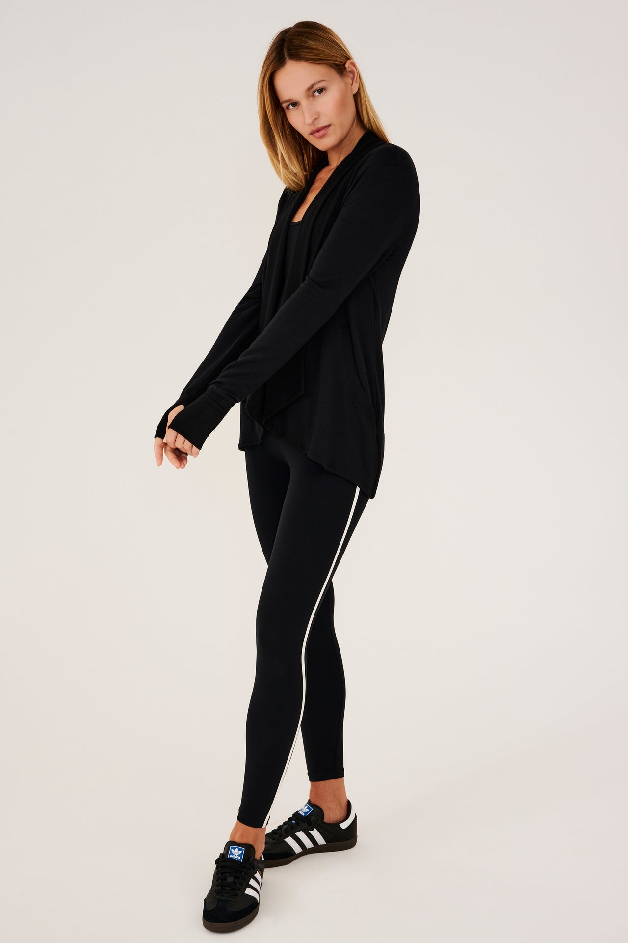 Full side view of girl wearing black long sleeve open front flowy sweater with thumb holes on sleeve cuff and black top with black leggings and black shoes