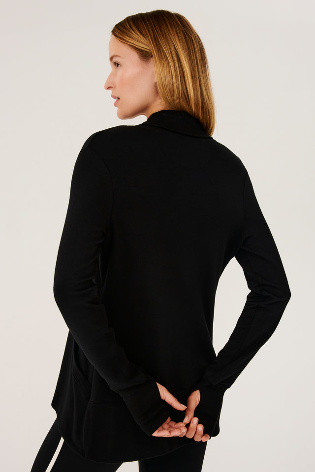 Back view of girl wearing black long sleeve open front flowy sweater with thumb holes on sleeve cuff and black top with black leggings