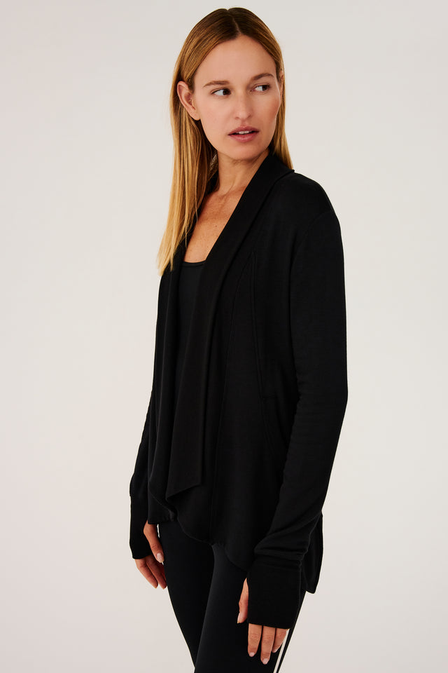 Side view of girl wearing black long sleeve open front flowy sweater with thumb holes on sleeve cuff and black top with black leggings