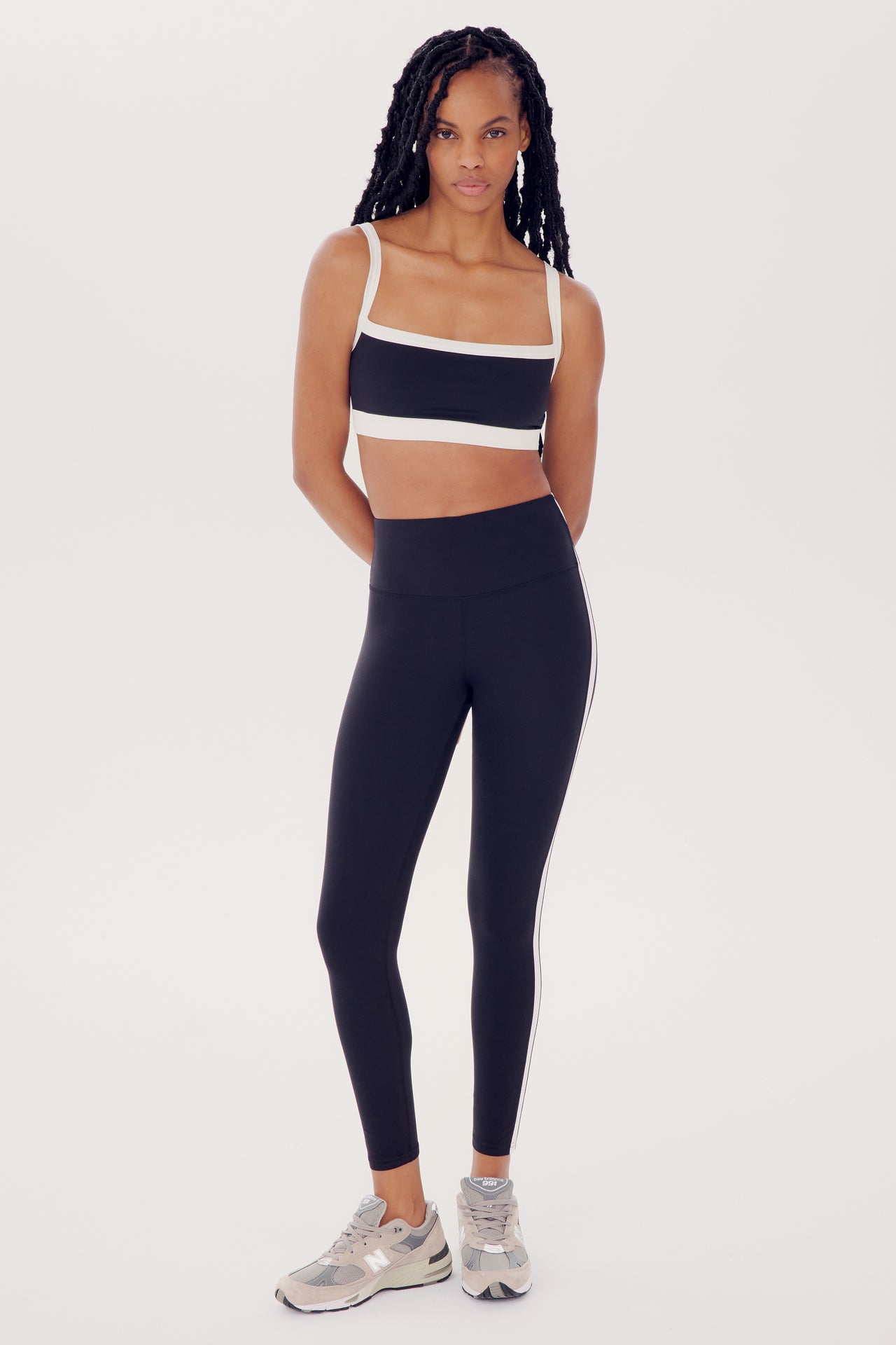 A woman in athletic wear, consisting of a black and white Monah Rigor Bra and leggings from SPLITS59, stands facing the camera with her hands by her sides.