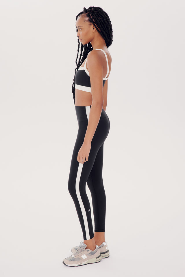 A woman in sportswear, including a black Monah Rigor Bra and leggings with white stripes, stands in profile on a white background.