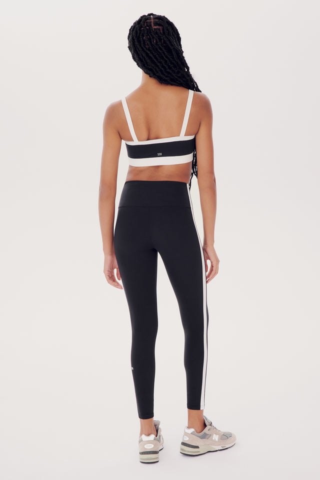 Rear view of a woman wearing sporty black leggings and a white Monah Rigor Bra by SPLITS59, standing against a white background.