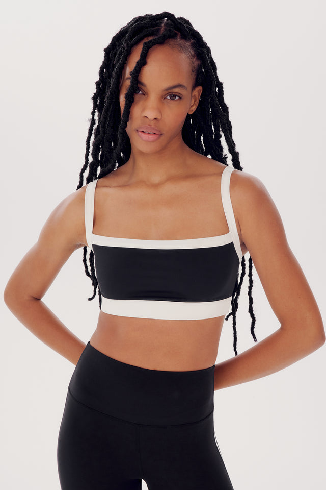 A woman in a black and beige SPLITS59 Monah Rigor Bra and leggings stands confidently, hands on hips, with long braided hair.