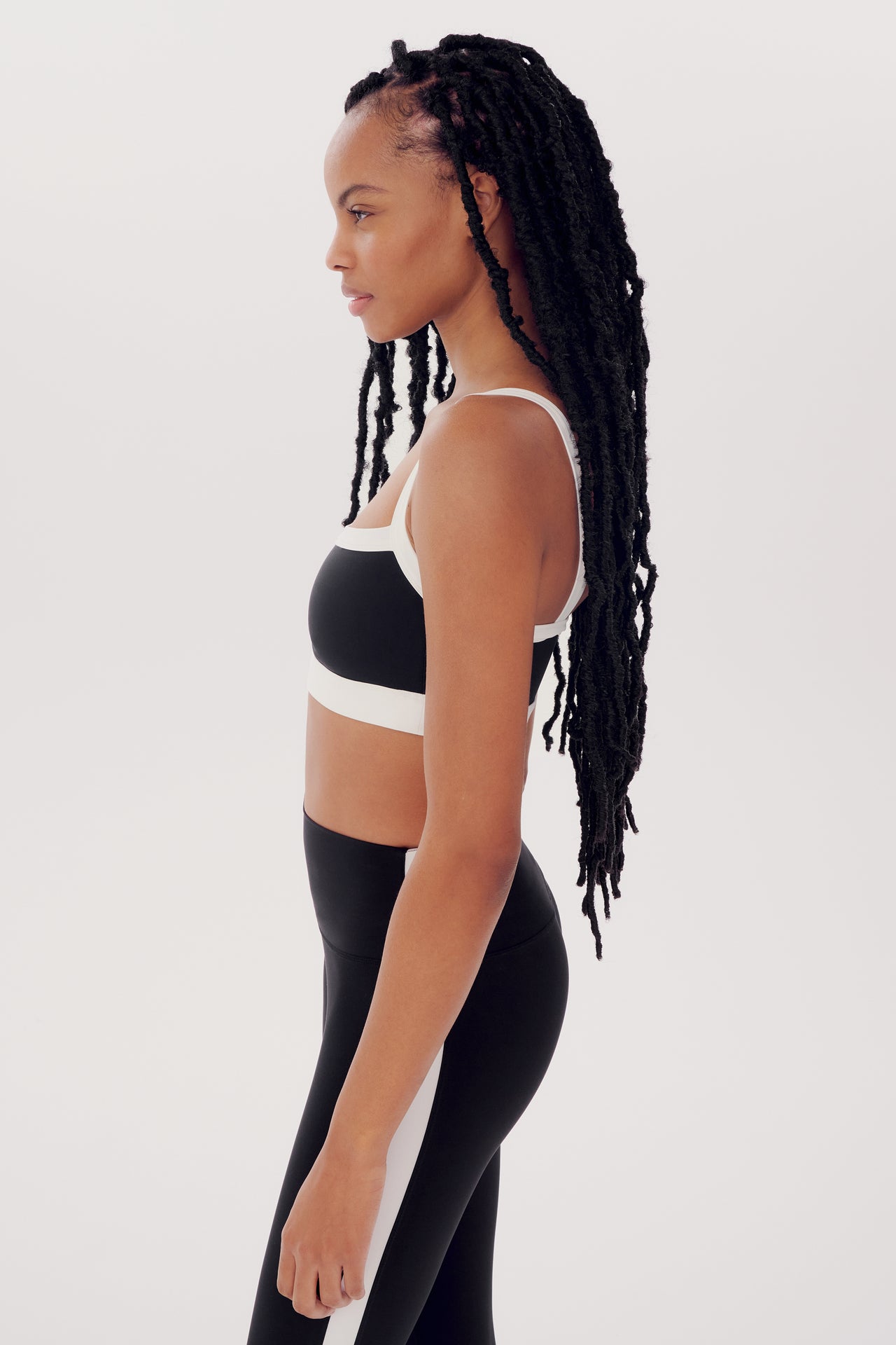 A woman in a black and white Monah Rigor Bra by SPLITS59 sports outfit stands in profile against a white background, with long braided hair.