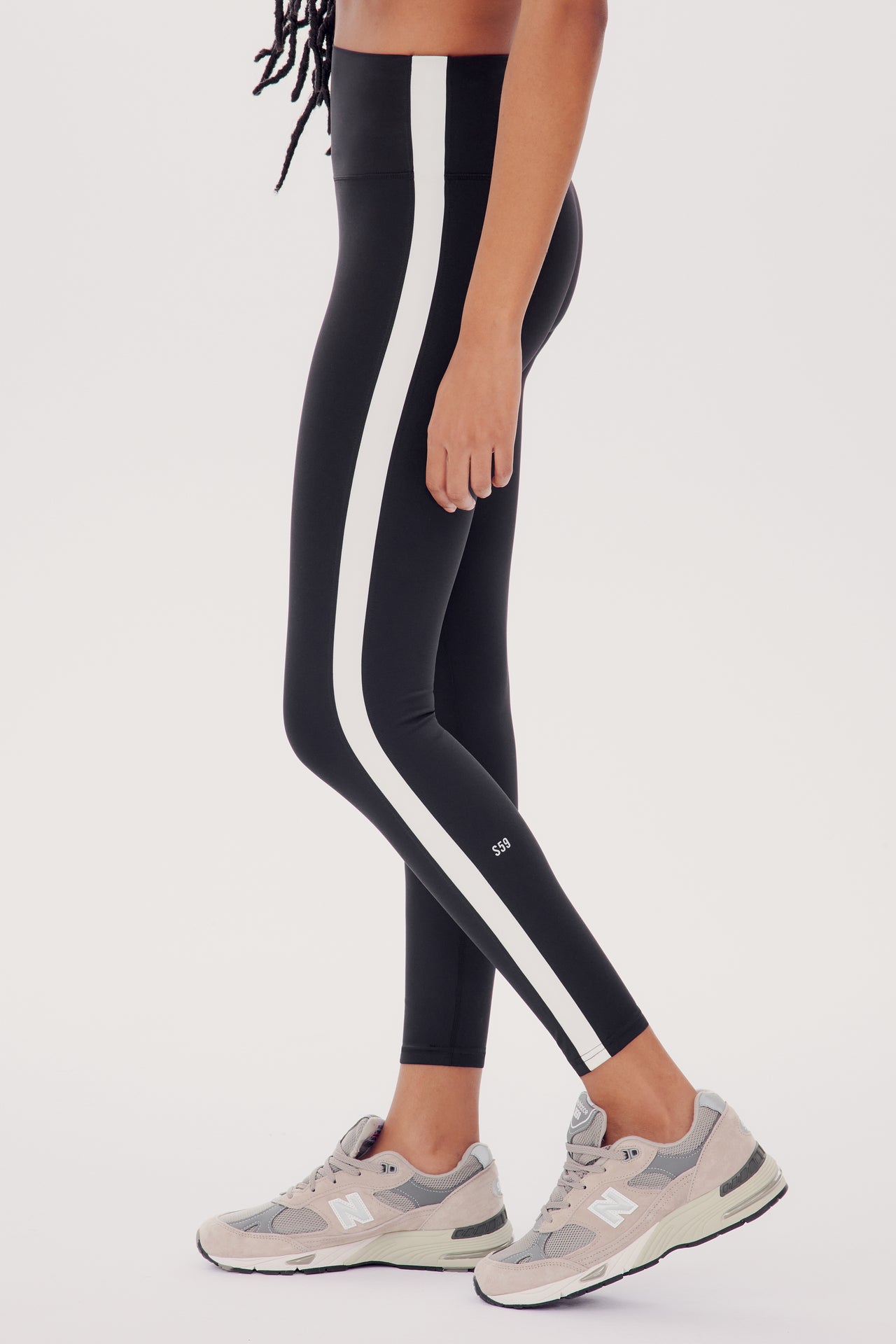 Woman wearing SPLITS59 Clare High Waist Rigor 7/8 leggings in Black/White with gray sneakers, standing against a white background, side view focusing on the legs.