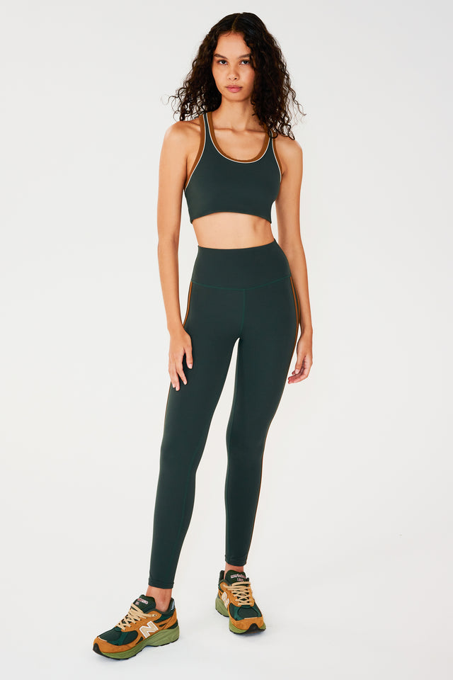 Full front view of girl wearing dark green leggings with two thin brown stripes down the side and a dark green sports bra with multi colored shoes
