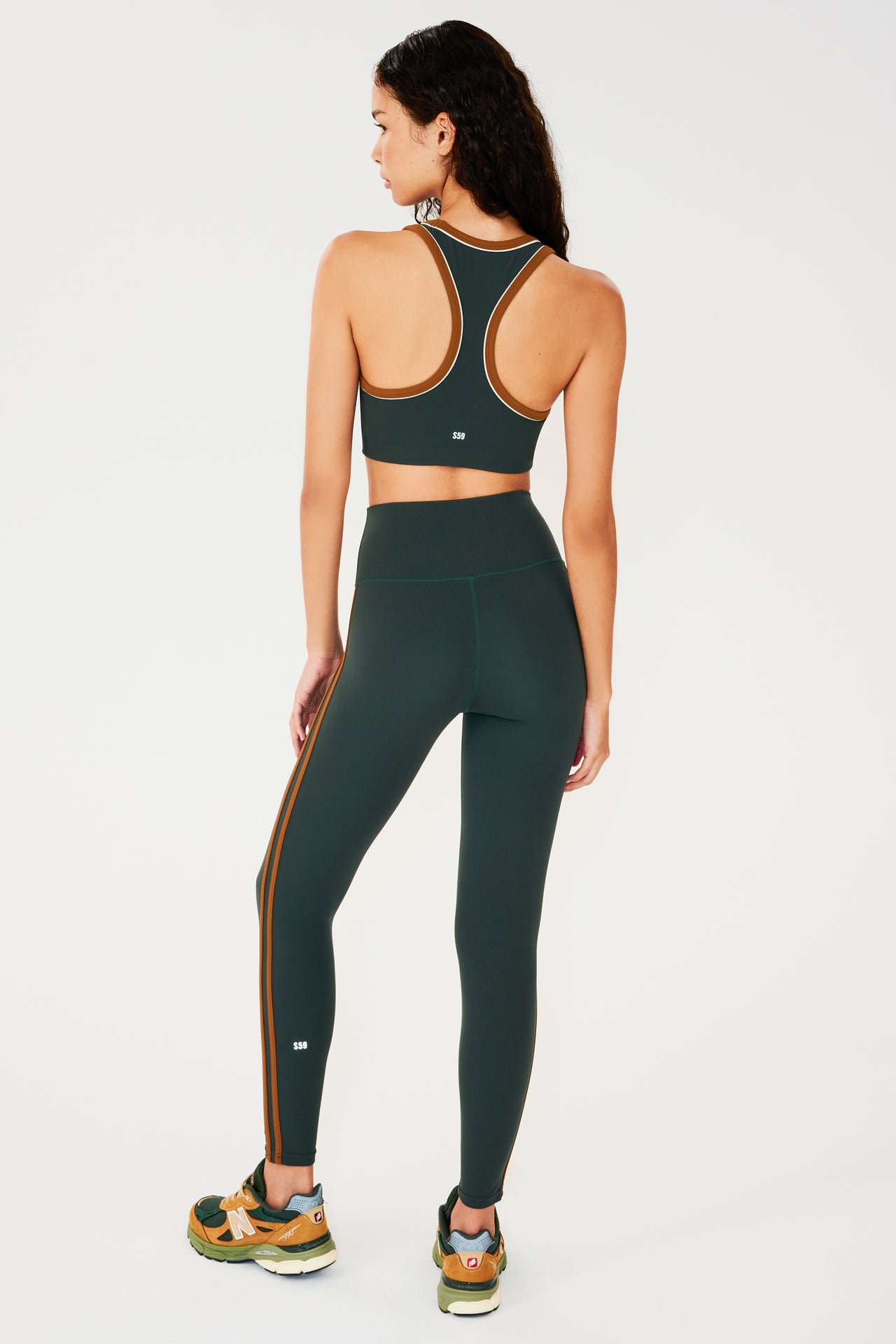 Full back view of girl wearing dark green leggings with two thin brown stripes down the side and a dark green sports bra with multi colored shoes