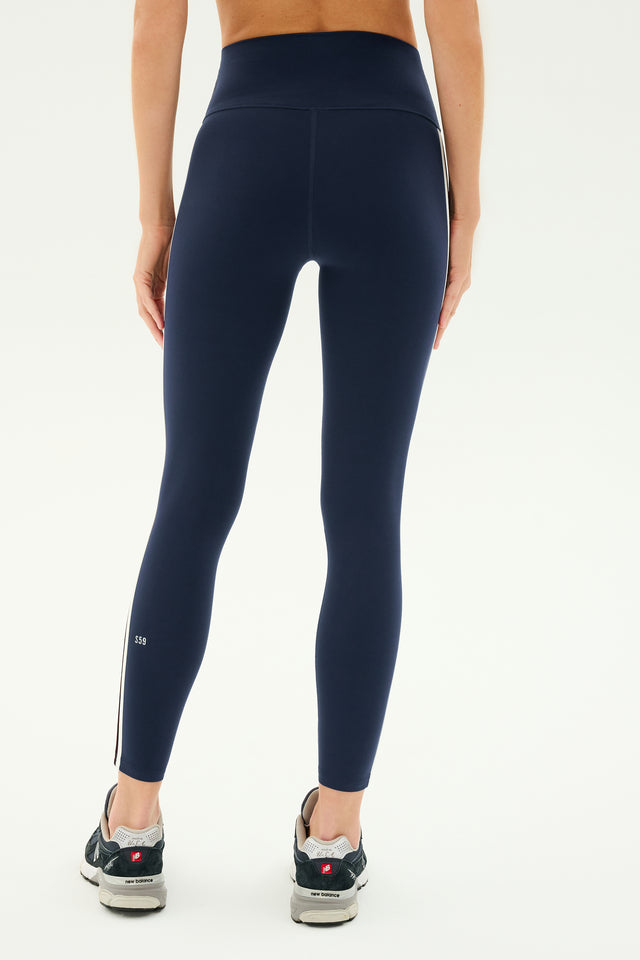 A person wearing SPLITS59 Ella High Waist Airweight 7/8 leggings in Indigo/Dark Cherry and gray sneakers is shown from the back, standing against a plain white background.