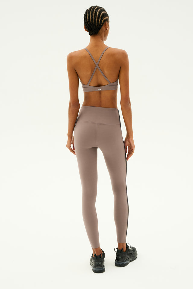 A person wearing a sports bra, Clare High Waist Rigor 7/8 leggings by SPLITS59 in Lentil/Black, and sneakers stands facing away against a plain background.