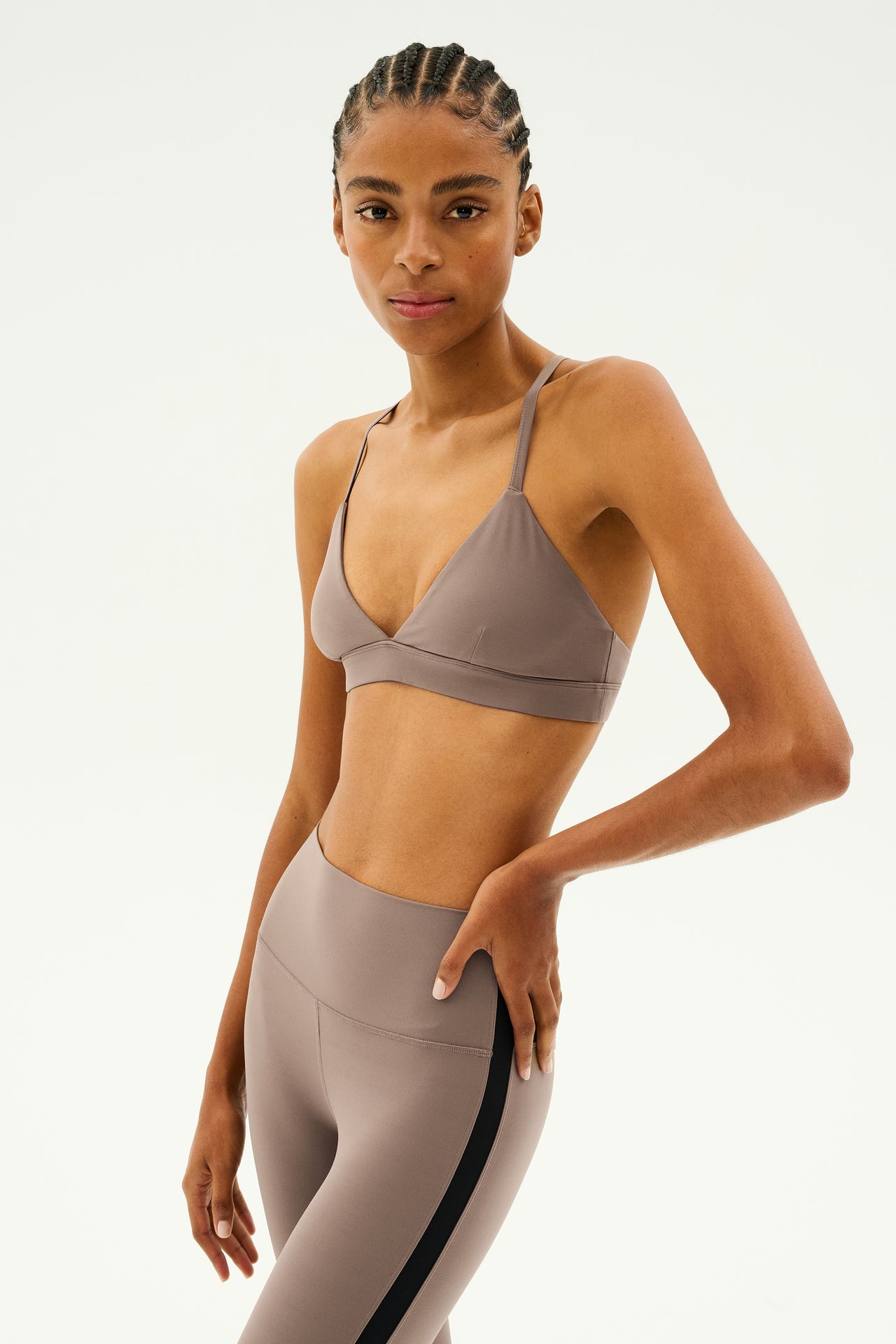 A person with braided hair wears a matching taupe SPLITS59 Theo Rigor Bra in Lentil and leggings made from ultra luxe Rigor fabric, posing against a plain background.