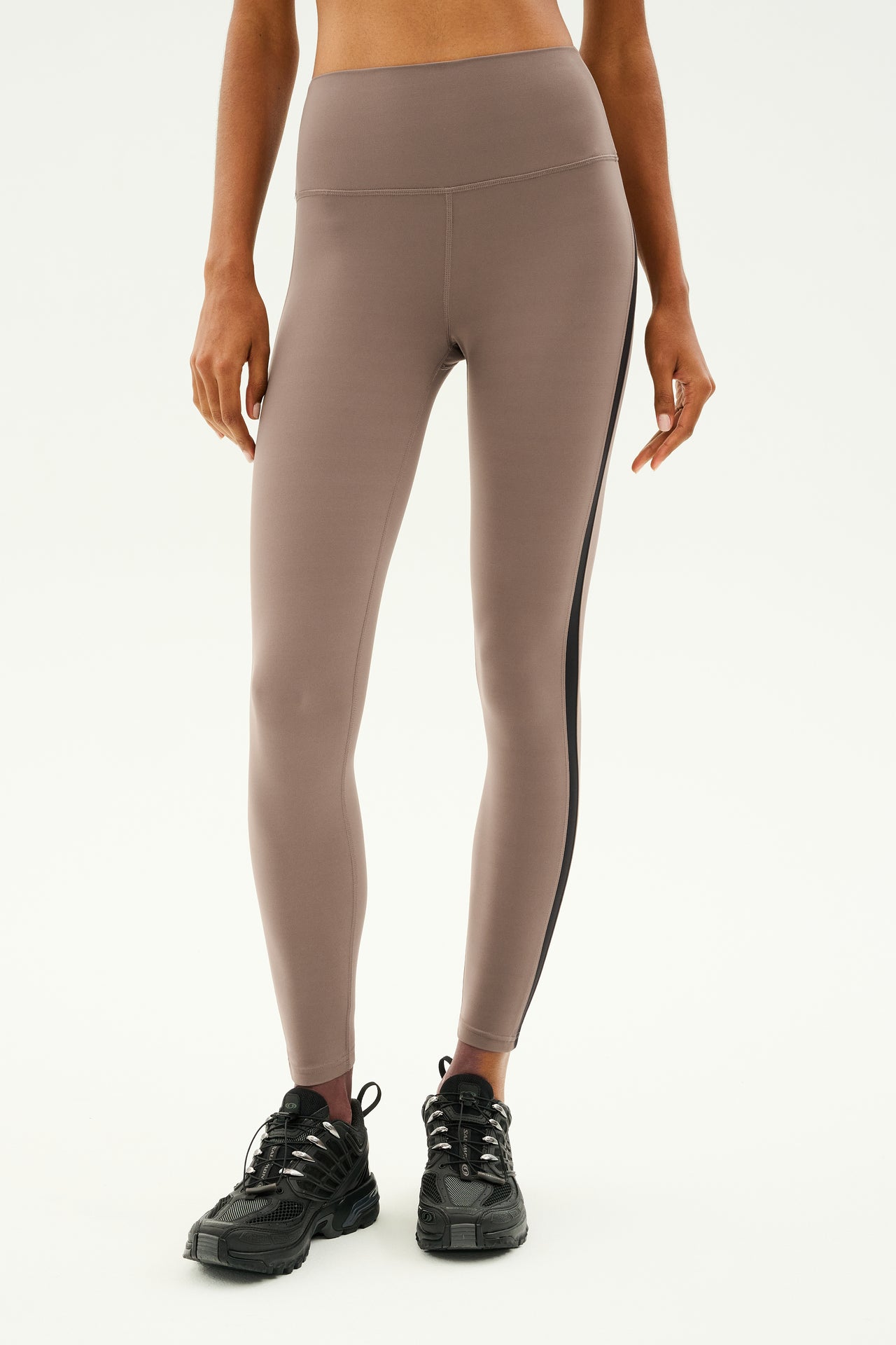 A person wearing SPLITS59 Clare High Waist 7/8 leggings in Lentil/Black, crafted from high-performance Rigor fabric, along with black sneakers stands against a light background.