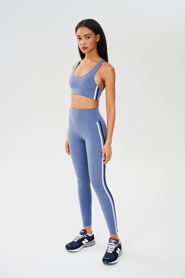 Full side view of girl wearing light blue leggings with thin white and black stripes down the side and a light blue sports bra with dark blue shoes