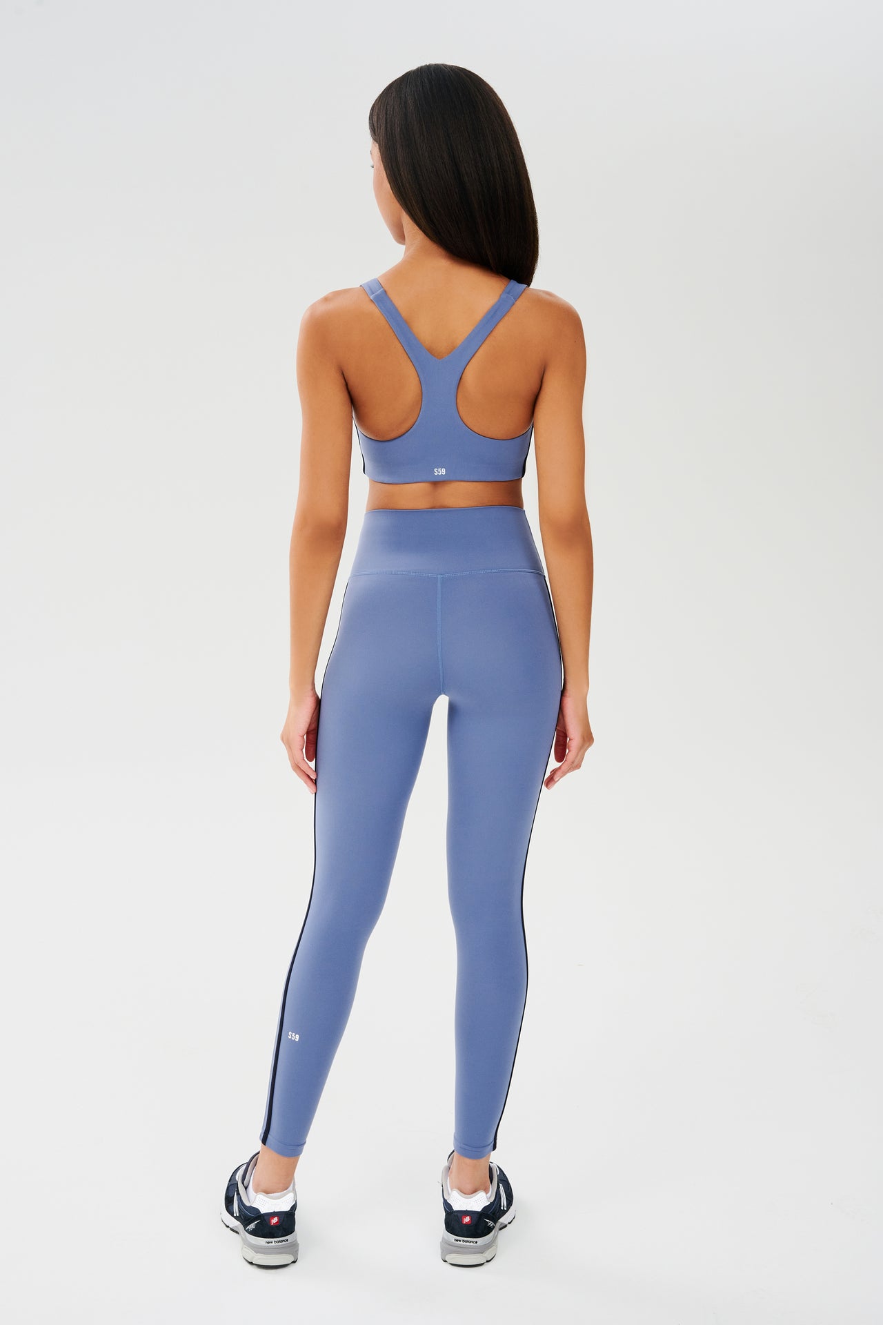 Full back view of girl wearing light blue leggings with thin white and black stripes down the side and a light blue sports bra with dark blue shoes