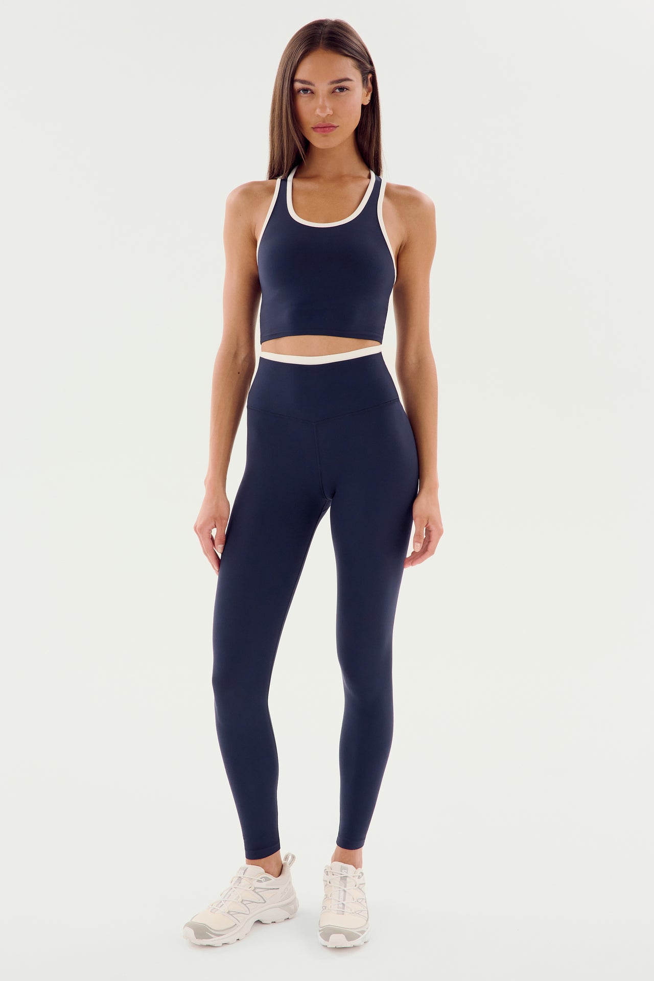 Woman standing in a fitness outfit consisting of a navy sports bra and matching Dual High Waist Airweight 7/8 - Indigo/White leggings from SPLITS59 made from ultra luxe fabric, paired with white sneakers. She has long, straight hair and poses against a plain white background, ready for her hot yoga session.