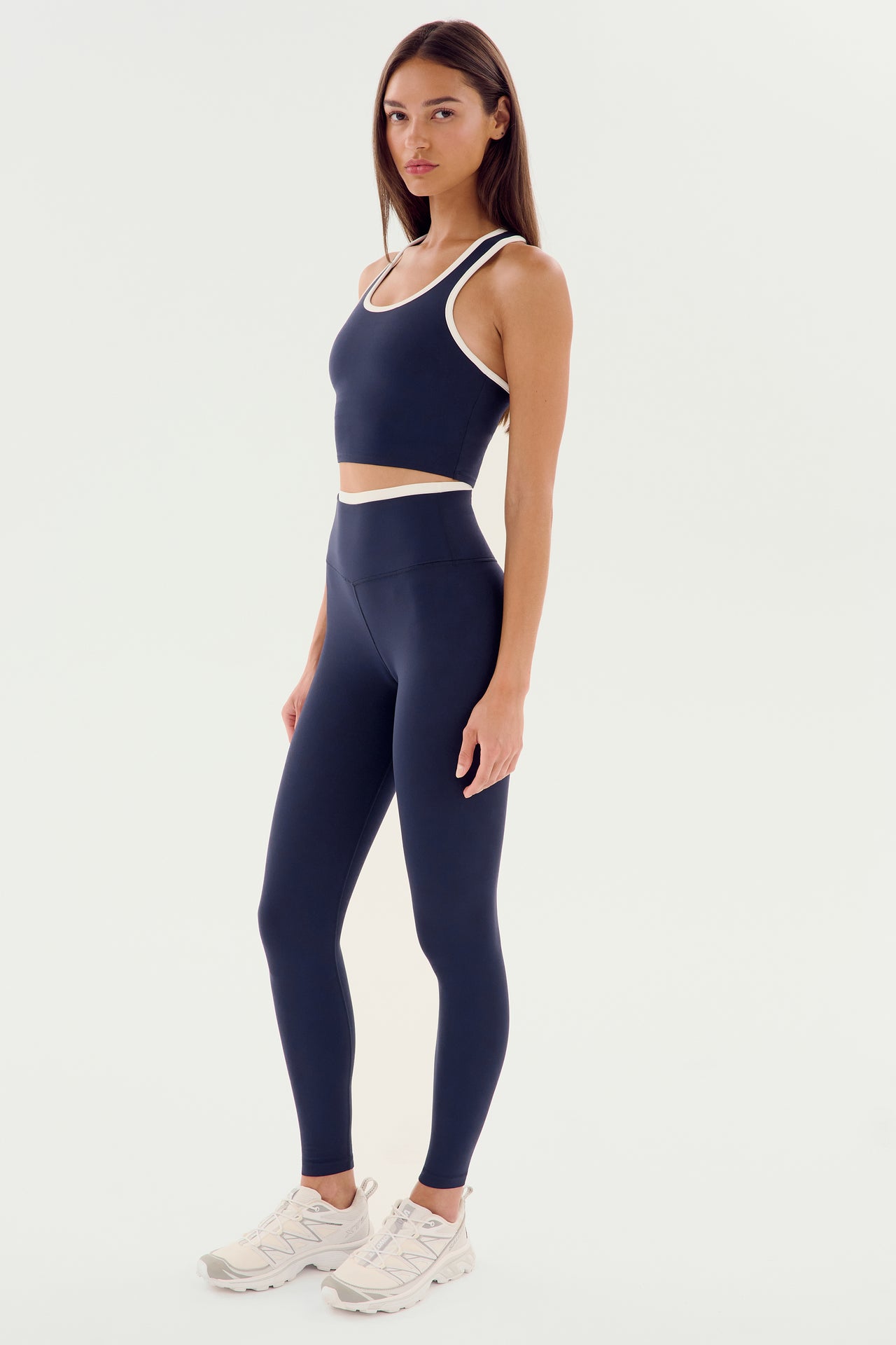 A person is standing and posing in a dark blue athletic outfit, consisting of a sleeveless top and matching Dual High Waist Airweight 7/8 - Indigo/White from SPLITS59, with white sneakers. The background is plain white.