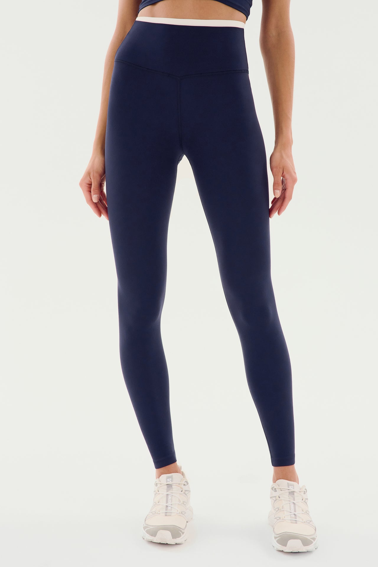 A person wearing SPLITS59 Dual High Waist Airweight 7/8 - Indigo/White leggings and white sneakers stands against a plain background.