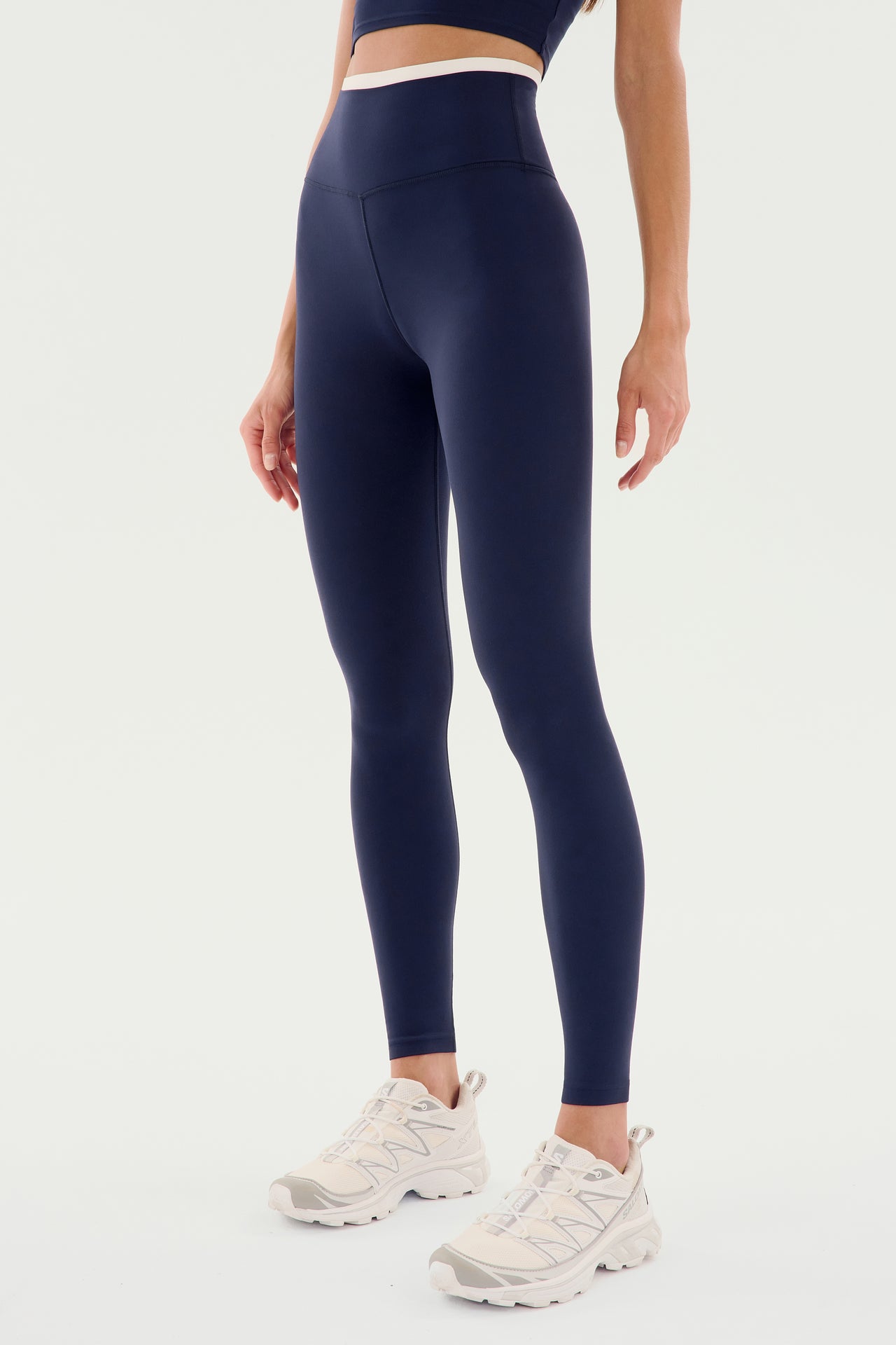 A person is wearing SPLITS59 Dual High Waist Airweight 7/8 - Indigo/White, perfect for Pilates, and white athletic shoes against a plain background. Only the bottom half of the body is visible.