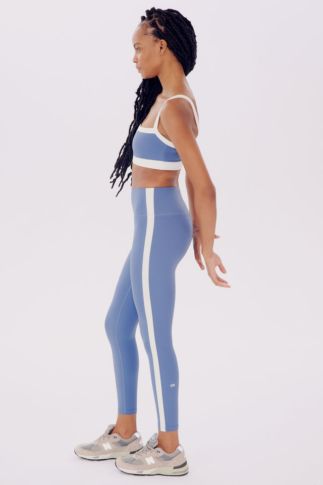 Side profile of a woman in blue SPLITS59 Monah Rigor Bra and sneakers, standing against a white background.