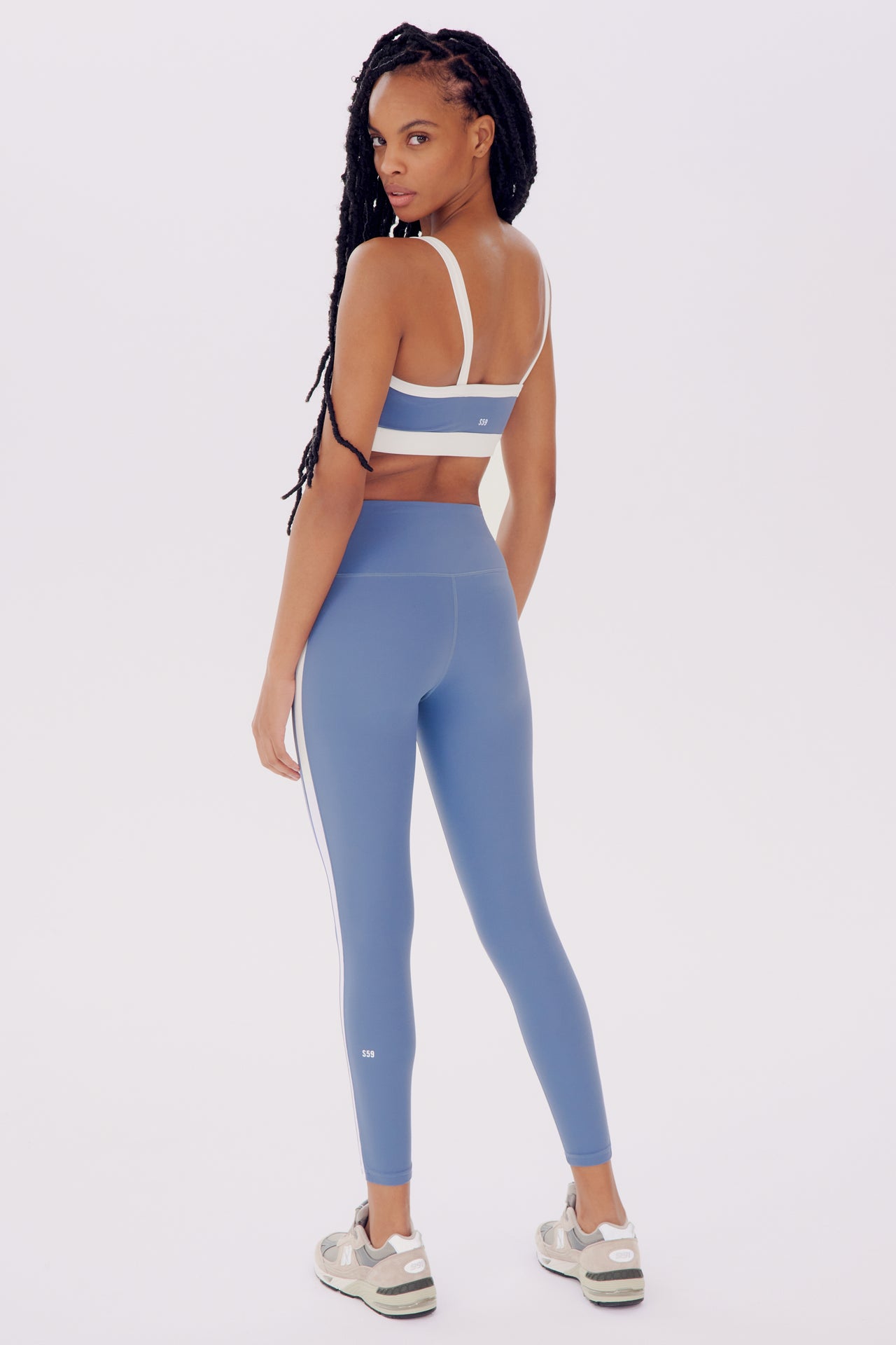 A woman in SPLITS59's Clare High Waist Rigor 7/8 - Steel Blue/White sports bra and high waist leggings turns her head to look over her shoulder, posing against a white background.