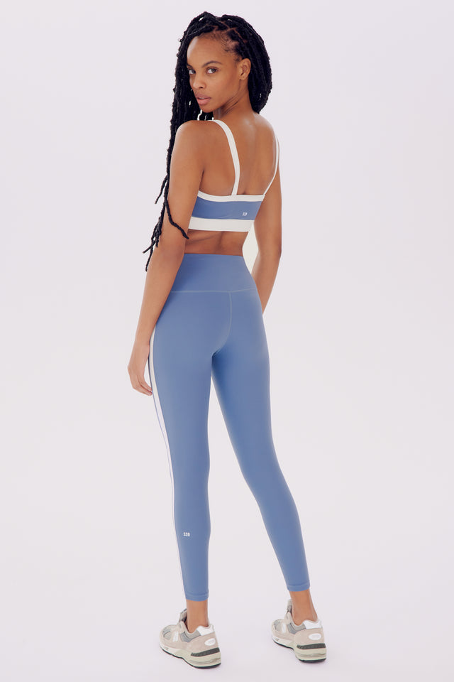 A woman wearing a SPLITS59 Monah Rigor Bra - Steel Blue/White and leggings, looking over her shoulder, against a white background.