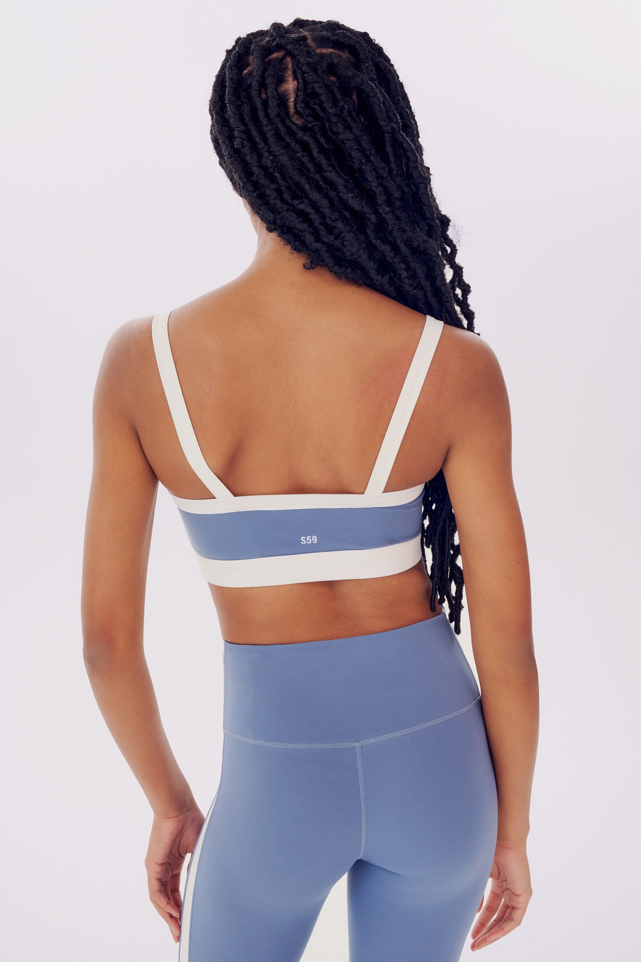 Rear view of a woman with braided hair wearing a SPLITS59 Monah Rigor Bra - Steel Blue/White and blue leggings standing against a plain background.