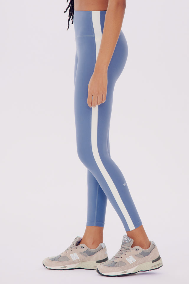 A person standing in profile wearing the SPLITS59 Clare High Waist Rigor 7/8 leggings in Steel Blue/White and gray sneakers, focusing on the legs from waist to feet.