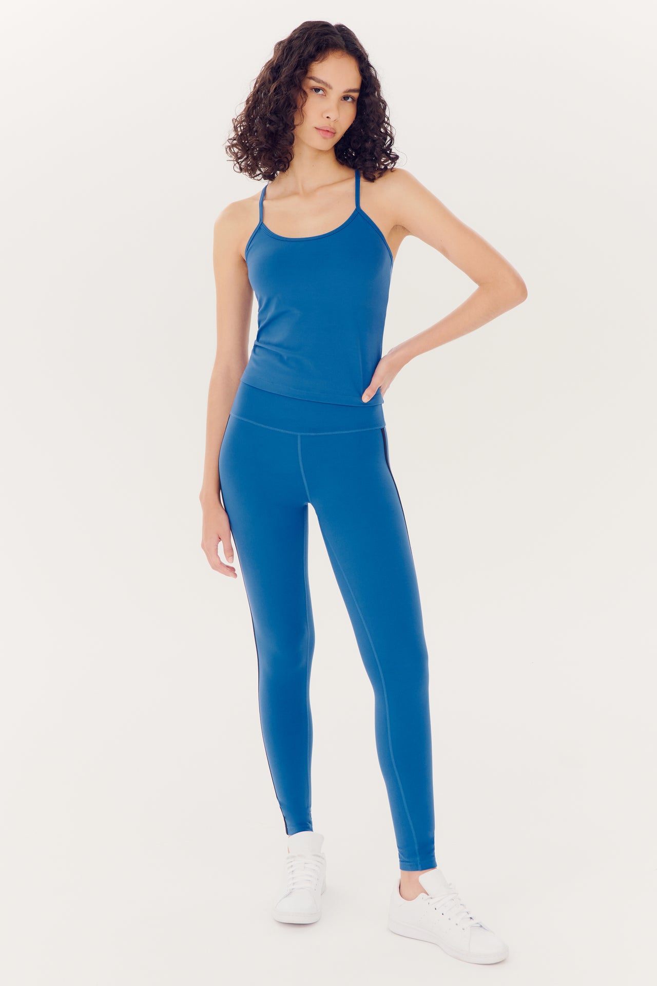 A woman wearing SPLITS59's Ella High Waist Airweight 7/8 leggings in Stone Blue/Indigo and a blue tank top stands against a white background, providing an accurate response.