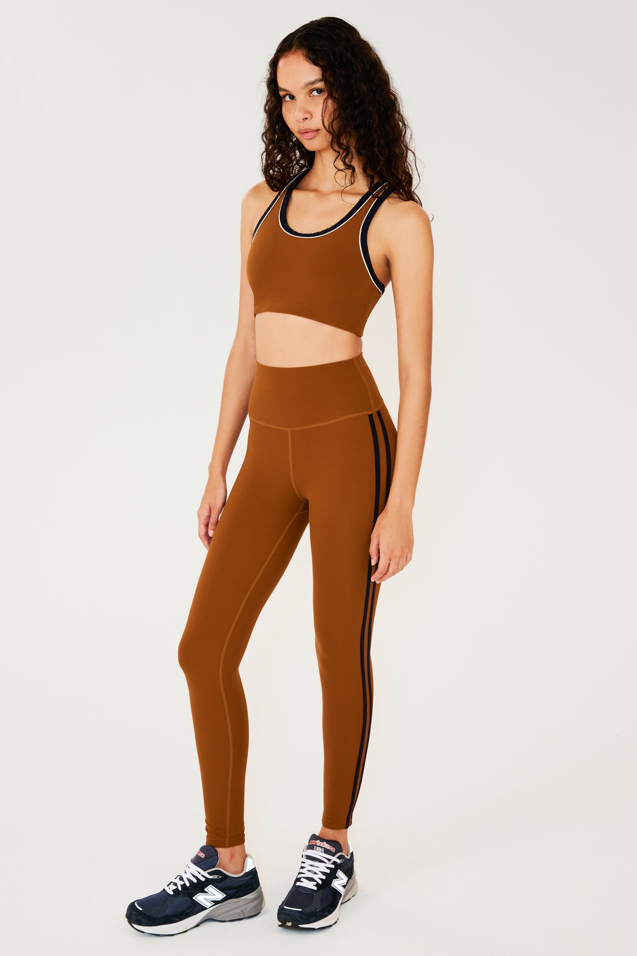 Full side view of girl wearing reddish brown leggings with two thin black stripes down the side and a reddish brown sports bra with dark blue shoes