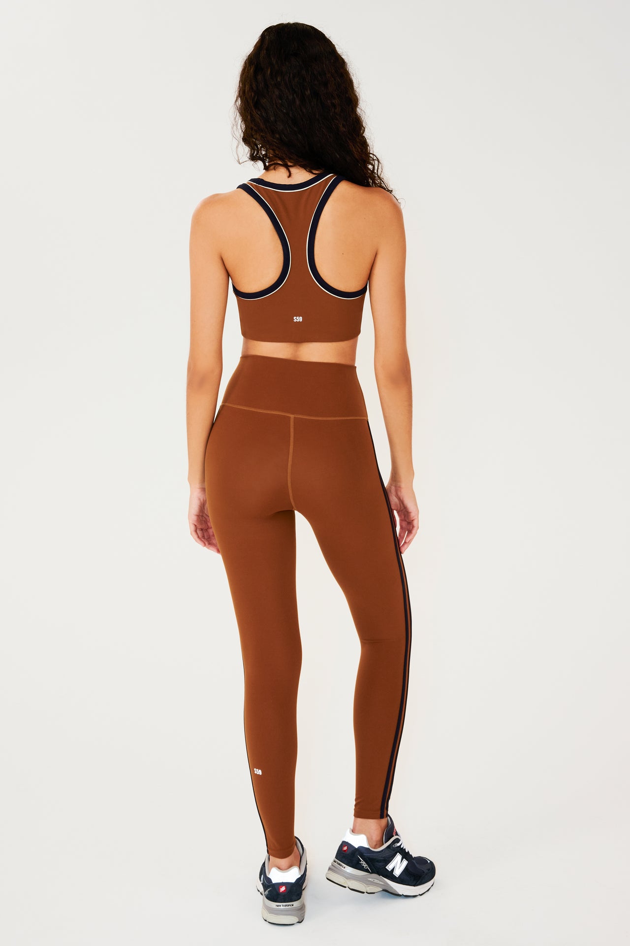 Full back view of girl wearing reddish brown leggings with two thin black stripes down the side and a reddish brown sports bra with dark blue shoes