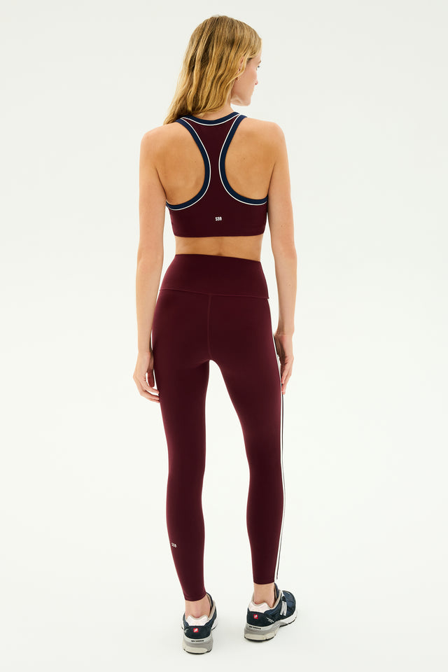 A woman, with her long blonde hair cascading down her back, is standing with her back to the camera, dressed in the Ella High Waist Airweight 7/8 leggings in Dark Cherry/Indigo from SPLITS59. The maroon sports bra and matching high waist leggings feature sleek white stripes and are crafted from butter-soft fabric. She's also wearing black sneakers.
