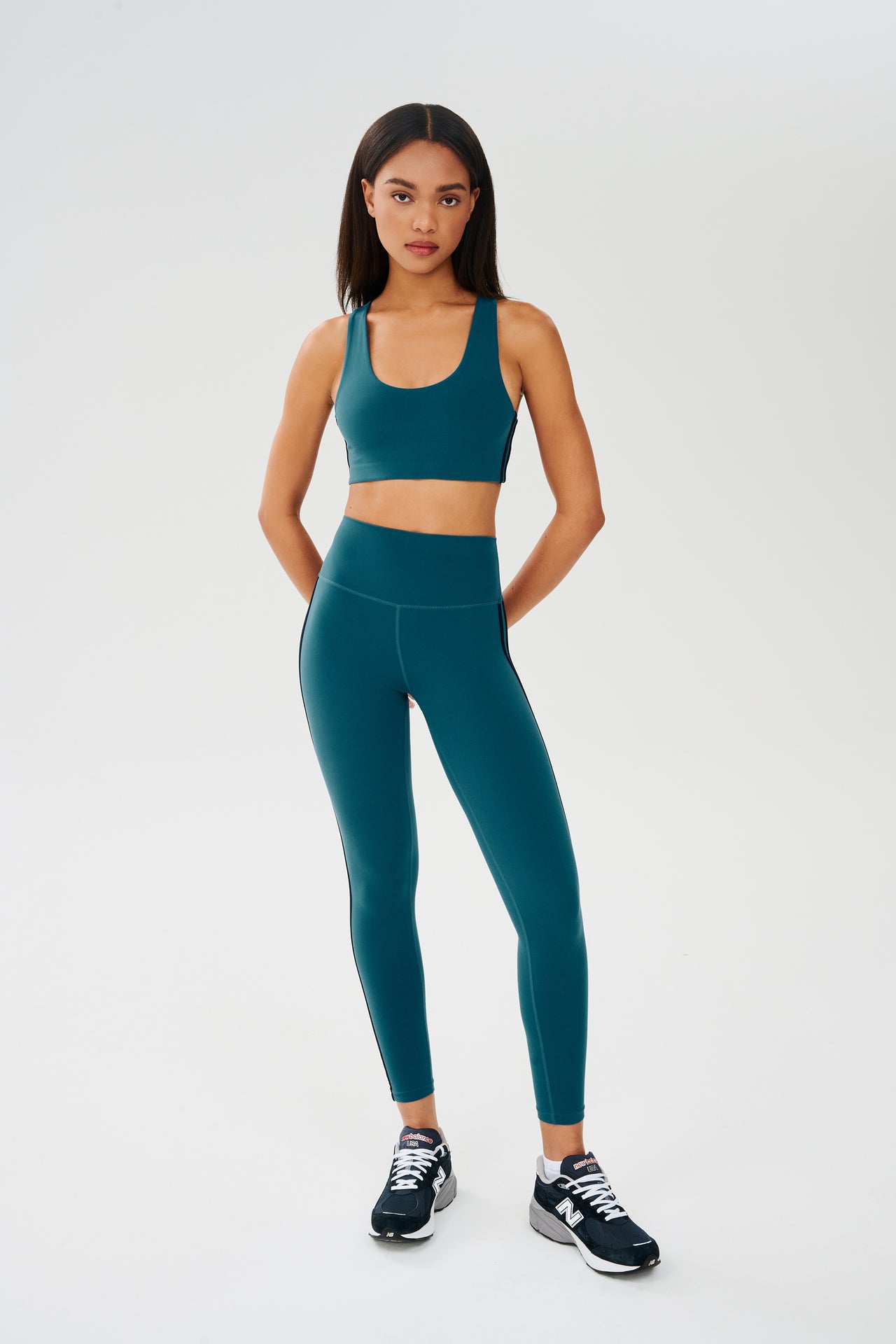 Full front view of girl wearing greenish blue sports bra with two thin black stripes down the side and greenish blue leggings with dark blue shoes