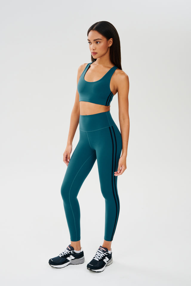Full side view of girl wearing greenish blue sports bra with two thin black stripes down the side and greenish blue leggings with dark blue shoes