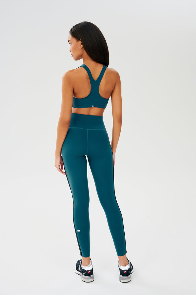 Full back view of girl wearing greenish blue sports bra with two thin black stripes down the side and greenish blue leggings with dark blue shoes
