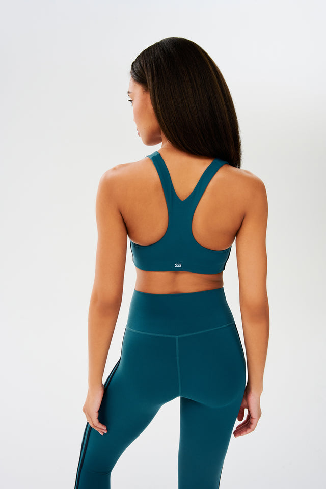 Back view of girl wearing greenish blue sports bra with two thin black stripes down the side and greenish blue leggings 