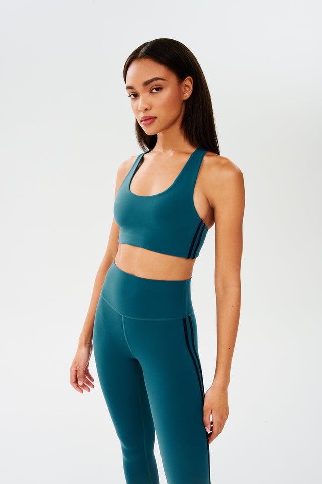 Side view of girl wearing greenish blue sports bra with two thin black stripes down the side and greenish blue leggings 